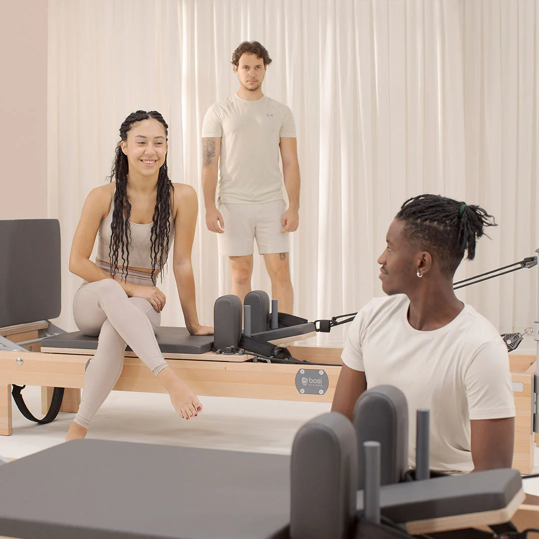 A man and woman are seated on a Pilates reformer, engaged in a workout session together.