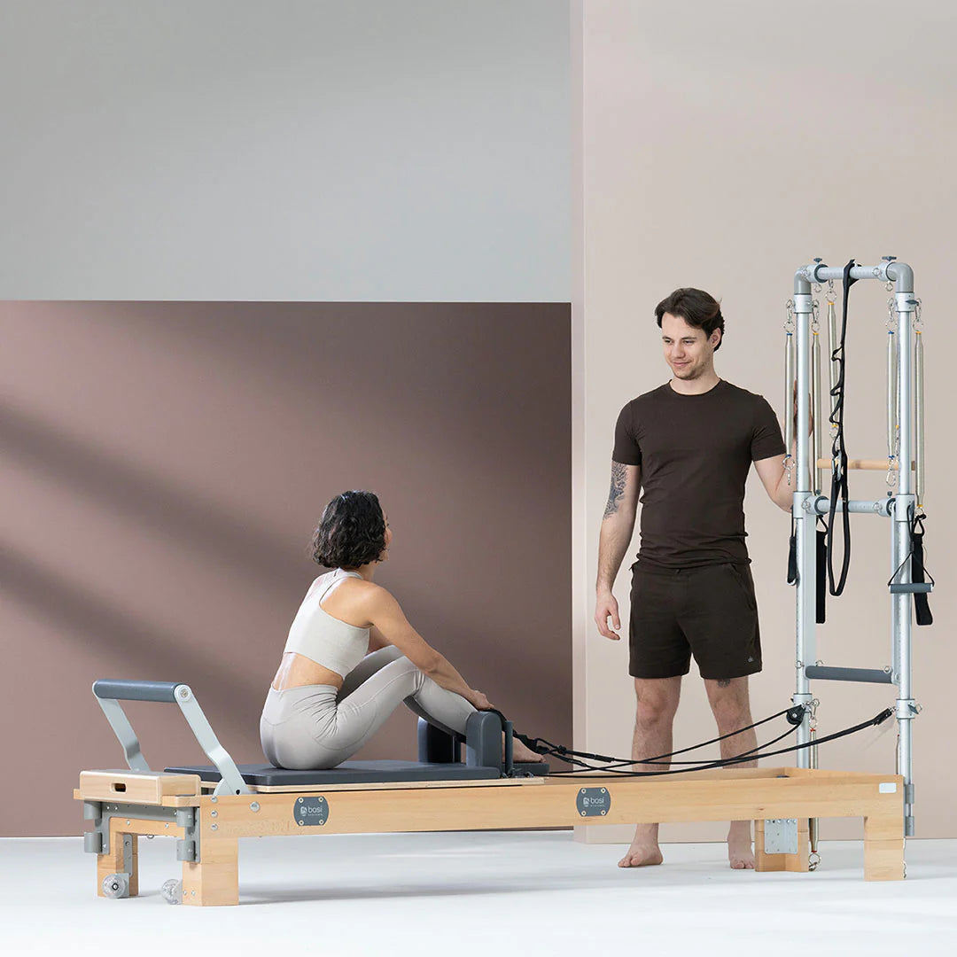 A man and woman stand beside a pilates machine, both appearing focused and ready for their workout session.