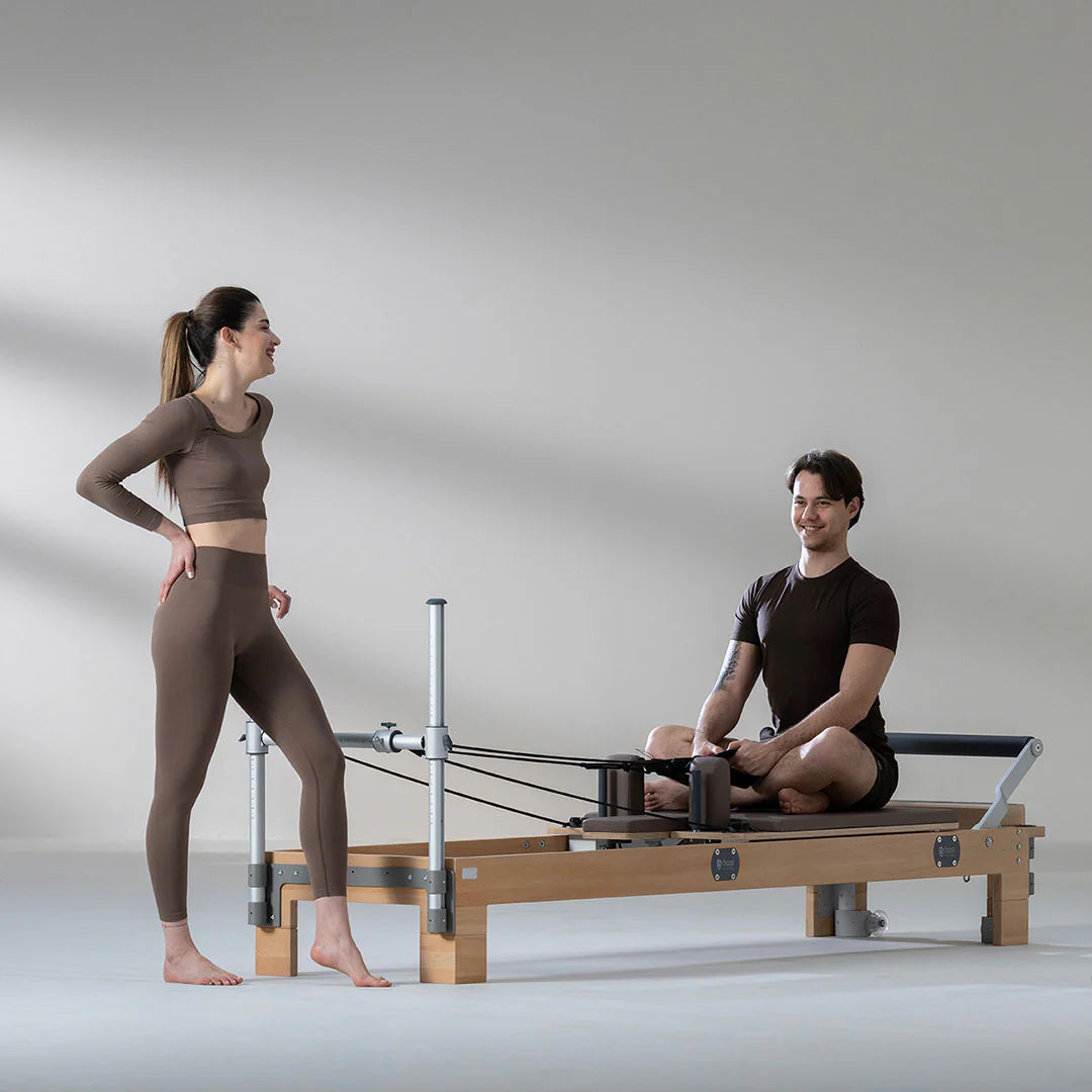 Man seated cross-legged on a Pilates Reformer, preparing for a workout, while a female instructor stands beside him smiling in a modern fitness studio setting.